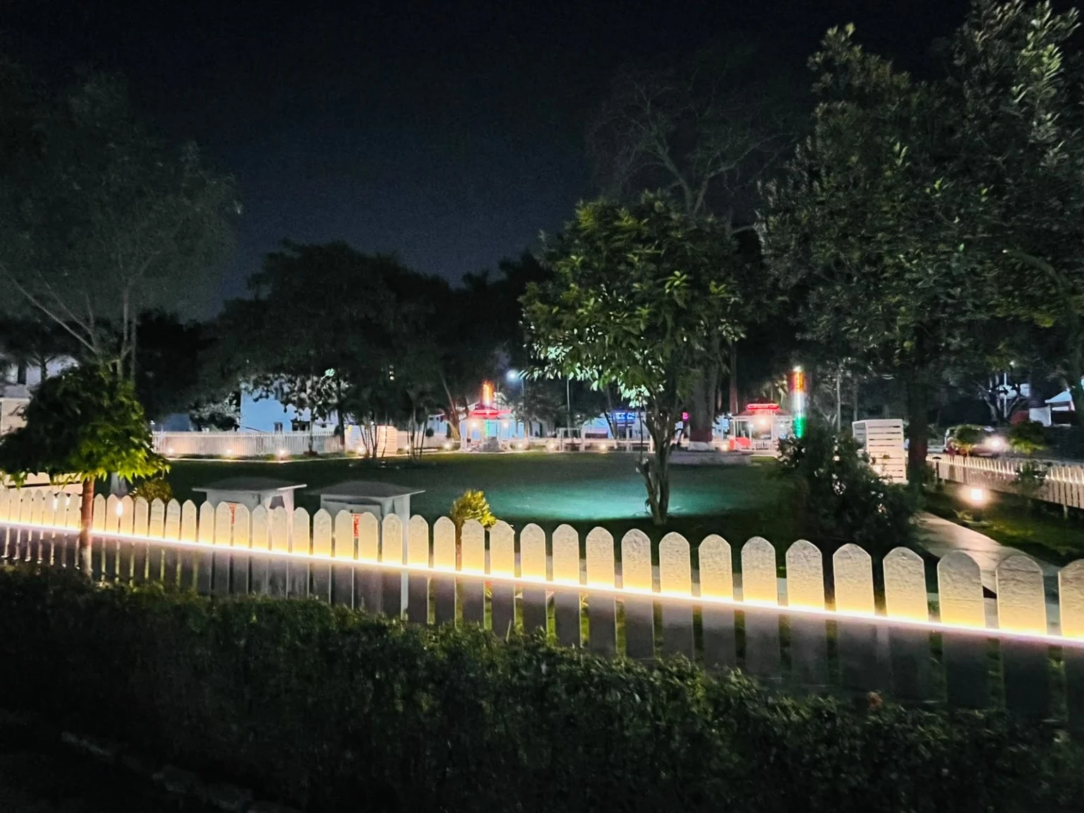 Light runs along a white garden fence beside a broad lawn at night.
